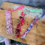 Three resin bookmarks with pressed flowers and tassels on a wooden board.