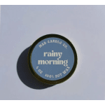 Round candle tin labeled "rainy morning" on a plain surface