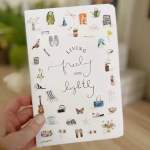 Hand holding a white notebook with small illustrated objects and the words "living freely and lightly"