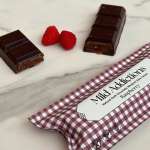 Dark chocolate raspberry bar pieces with two raspberries and a checked Mild Addictions wrapper on marble.