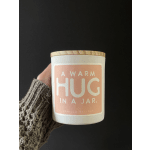 A white candle jar with a wooden lid and peach label reading "A Warm Hug in a Jar"