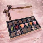 Open pink gift box of assorted square and heart-shaped chocolates with a brown ribbon bow.