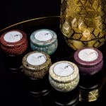 Assorted patterned candle jars with gold lids on a dark surface.
