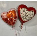 Two heart-shaped Valentine balloons with Spanish text against a wall