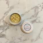 Two small tin candles on a marble surface, one open and one with a labeled lid