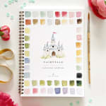 Watercolor workbook with a castle illustration, paint swatches, brush, and ribbon on a white surface