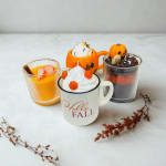 Three fall-themed candles in mugs and glass cups with pumpkin decorations