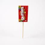 Skittles candy package on a wooden pick against a white background