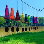 Row of multicolored glass bottle wind chimes hanging from a metal rod outdoors by a river
