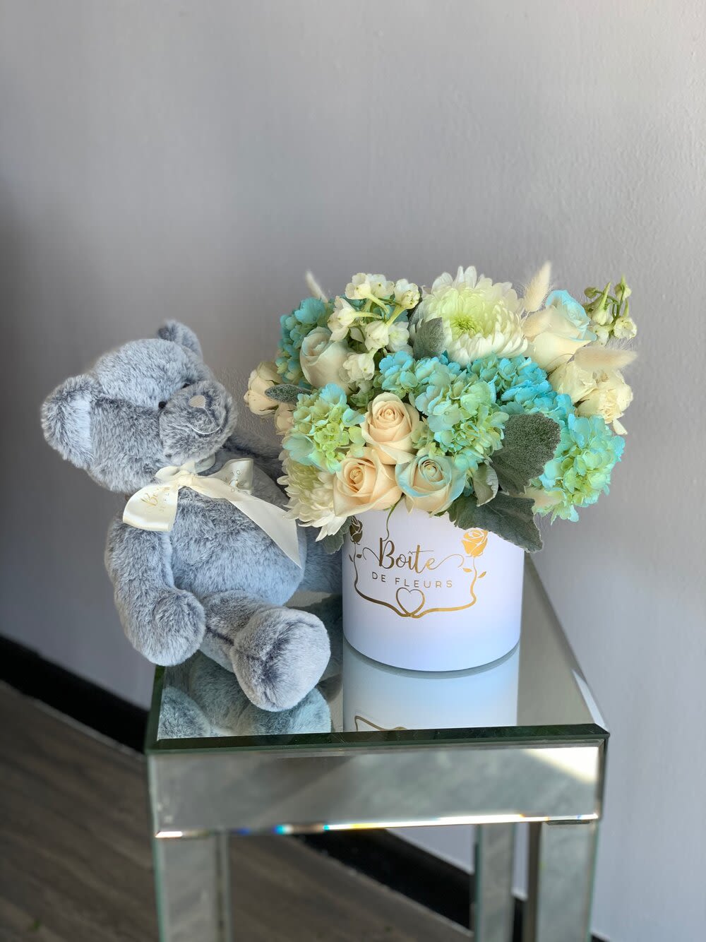 Blue teddy bear beside a pastel flower arrangement in a white box