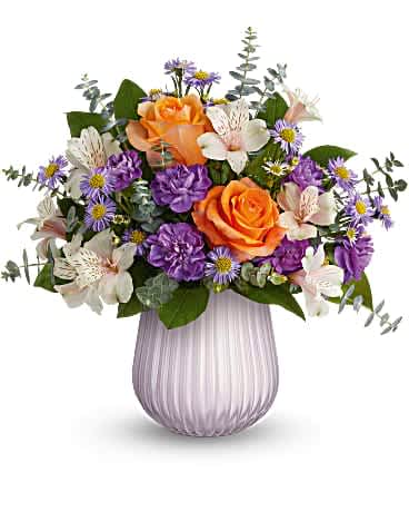 Arrangement of orange roses, purple carnations, asters, and white alstroemeria in a pale lavender ribbed vase