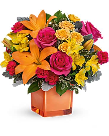 Bright arrangement of orange lilies, pink roses, and yellow blooms in an orange cube vase