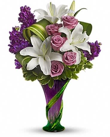 Tall arrangement of white lilies and pink roses with purple blooms in a purple and green glass vase