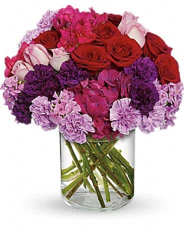 Compact bouquet of red and pink roses with purple and lavender flowers in a clear glass vase.