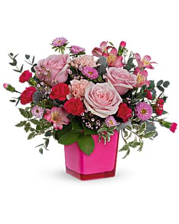 Pink and red roses, carnations, and mixed flowers arranged in a bright pink square vase
