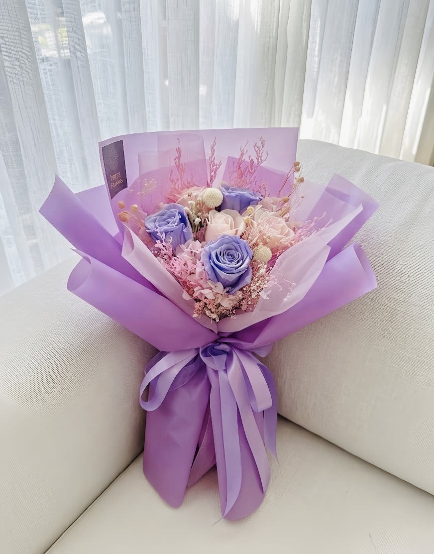 Lavender Dream - Preserved Rose Bouquet - A dreamlike preserved flower bouquet featuring 3 premium lavender-purple roses and 3 soft cream-pink roses. This enchanting arrangement is beautifully layered with pink preserved hydrangeas, white baby's breath, and delicate dried floral fillers. Elegantly wrapped in layered lavender and translucent frosted paper, it is finished with a matching purple satin ribbon—making it an enduring symbol of love and appreciation.