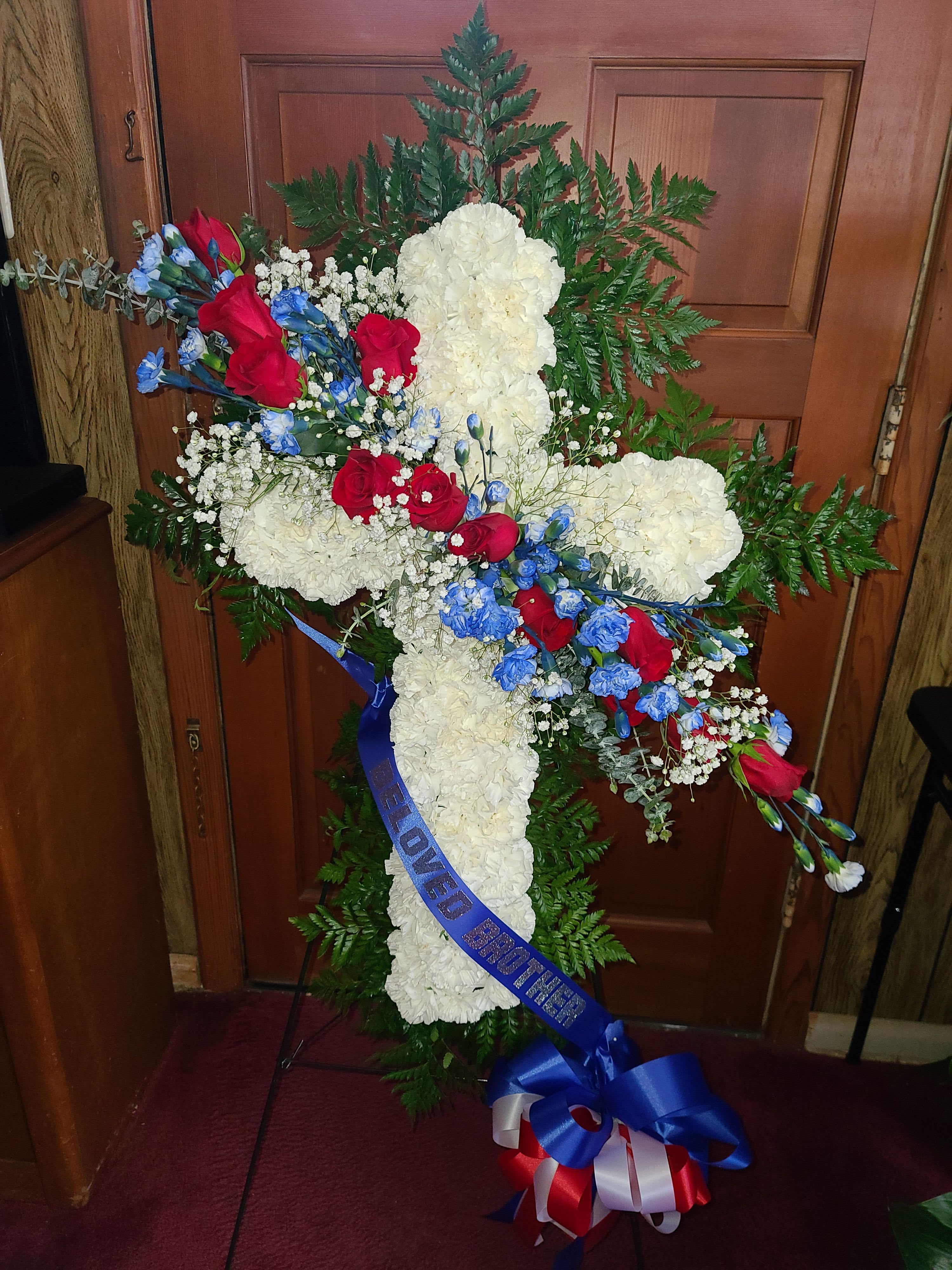 Cross-shaped floral tribute with white carnations, red roses, and blue flowers