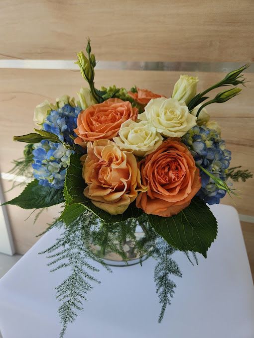 Compact arrangement of peach and cream roses with blue hydrangeas in a clear glass vase with feathery greenery.