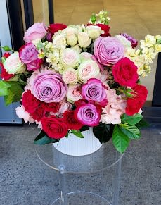 Round arrangement of pink, red, and white roses in a white hatbox-style container.