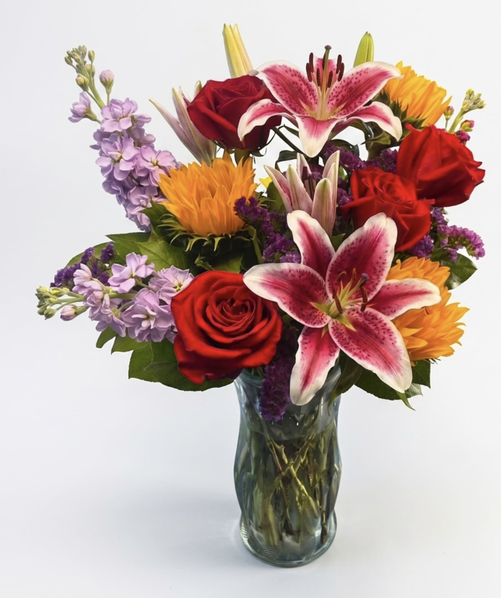 Golden Embrace for Mom  - Make this Mother’s Day unforgettable with Golden Embrace for Mom, a breathtaking floral arrangement designed to capture warmth, joy, and appreciation in every bloom. This vibrant design features cheerful sunflowers paired with fragrant stargazer lilies, premium roses, soft yellow stock, and rich purple statice—creating a perfectly balanced bouquet full of texture, color, and elegance. Thoughtfully arranged in a clear glass vase with lush greenery, this design offers both height and fullness, making it a standout centerpiece for any home. The combination of bold sunflowers and romantic roses symbolizes admiration and love, while the stargazer lilies add a luxurious touch and unforgettable fragrance. Whether you’re celebrating Mom, a grandmother, or someone who means the world to you, this arrangement delivers beauty and emotion in every detail.