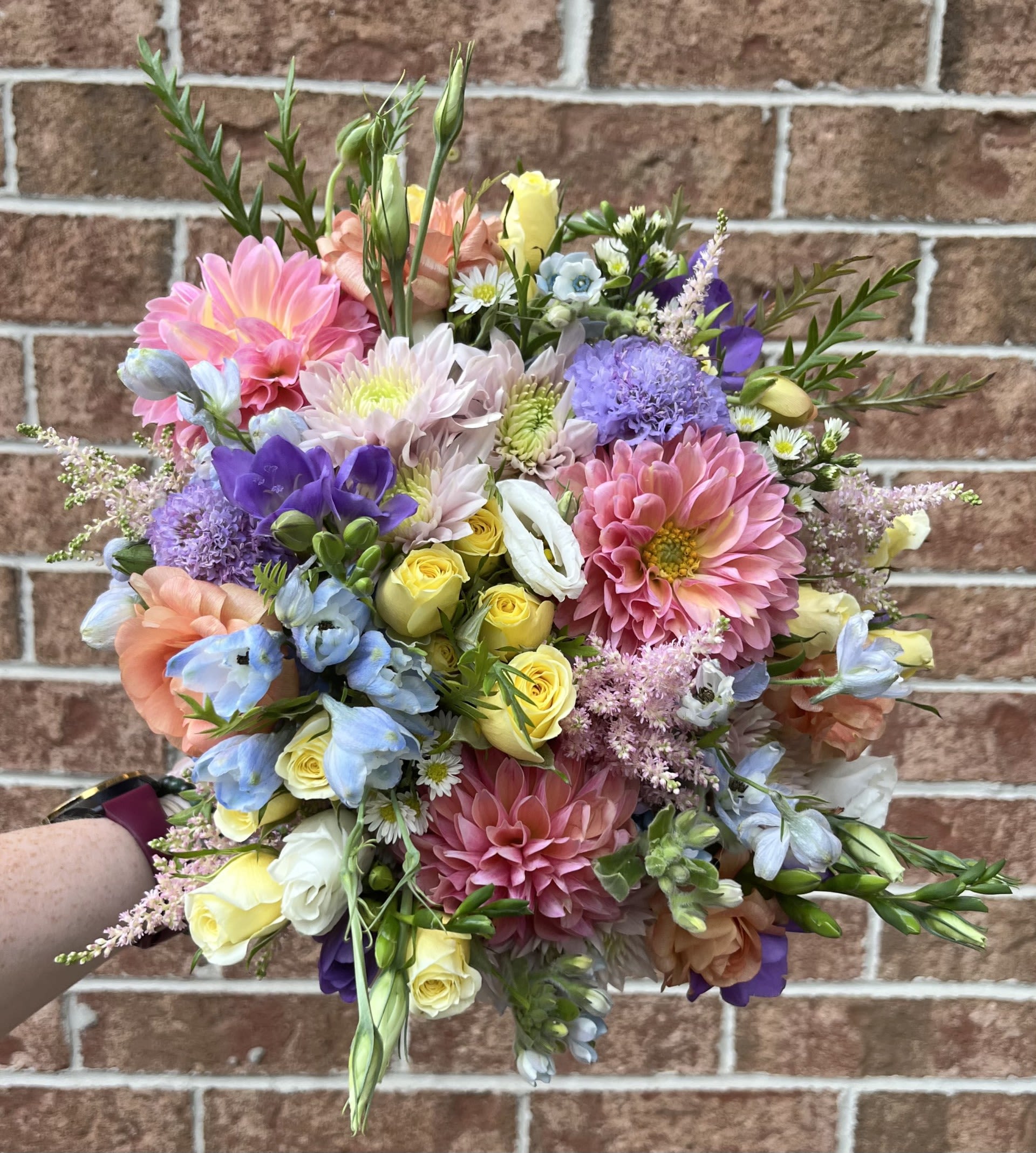 Spring Garden Bridal Bouquet by Chasing Petals - Step into a blooming daydream with this vibrant bouquet bursting with seasonal charm. Featuring a joyful mix of dahlias, roses, scabiosa, freesia, and delicate filler florals in pastel pinks, peaches, soft blues, and buttery yellows— this bouquet captures the whimsical beauty of an English garden in full bloom. Perfect for spring and summer brides who love a romantic, fresh-picked feel.