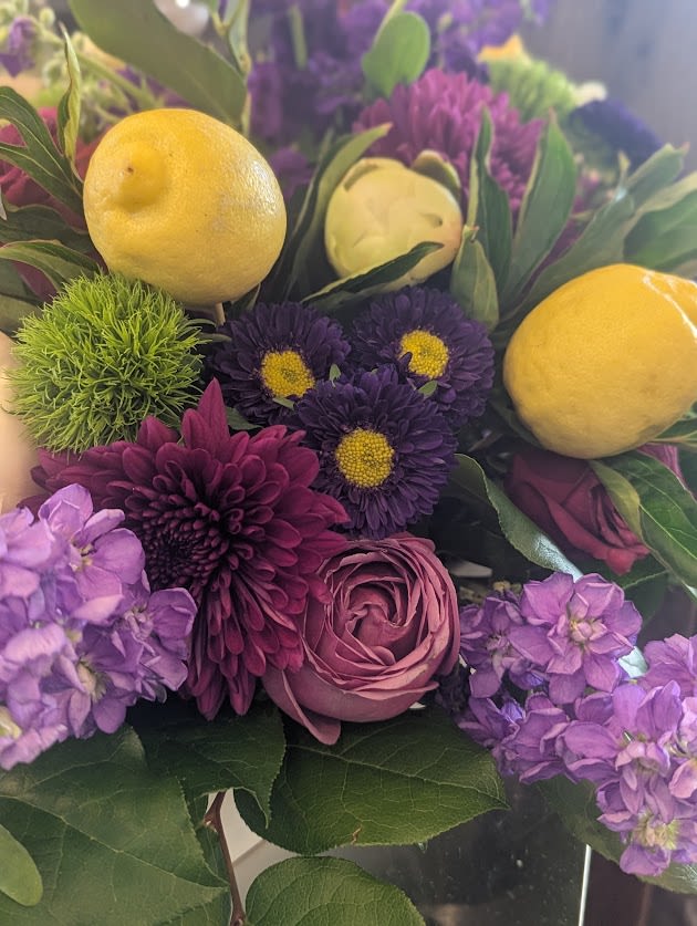 Arrangement of purple flowers with lemons and green pom blooms in a glass vase