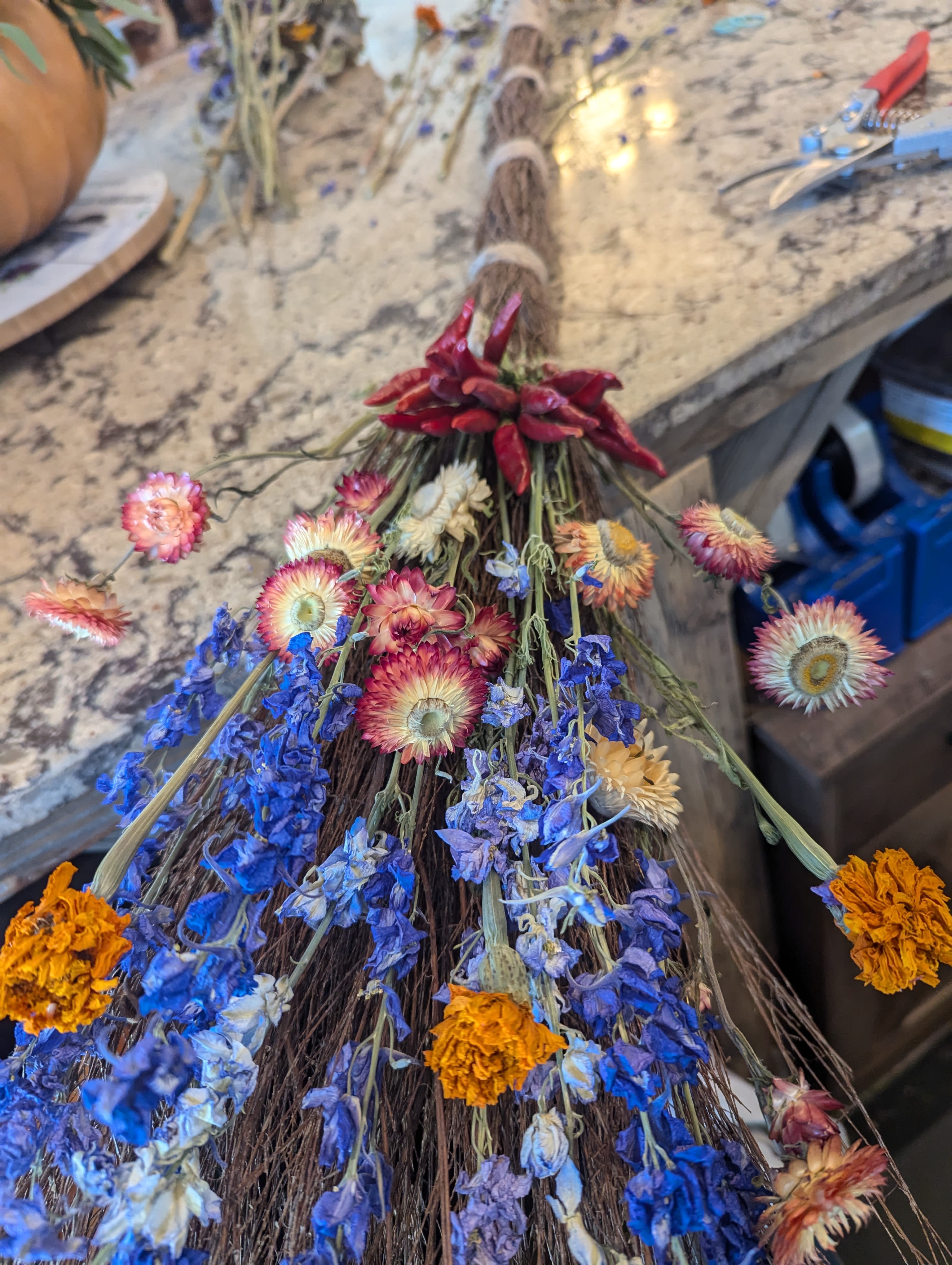 Rustic twig broom decorated with dried blue, orange, and cream flowers and red peppers.