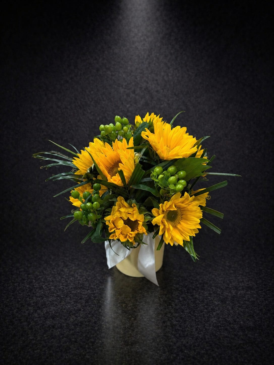 Golden Touch - A burst of sunny warmth-Golden Touch is a yellow sunflower arrangement that feels like a personal ray of light for birthdays, "just because" surprises, or a bright get-well gift. The compact, rounded design puts bold sunflower faces front and center so the person you're thinking of will instantly feel noticed and lifted. It also reads fresh for spring and summer, and its warm tones work beautifully into autumn and thank-you moments.  The bouquet pairs vivid yellow sunflowers with green berry-like accents and long, dark foliage for contrast and texture, all arranged in a light ceramic vase tied with a simple white ribbon. The ceramic keeps the look clean and gift-ready, while the close cluster of blooms creates a generous, cheerful silhouette that fits on a bedside table or kitchen counter.  Perfect for cheering someone after a rough week, saying thanks in a sunny way, or marking a birthday with casual elegance, this arrangement arrives ready to display. Care is simple: place in fresh water out of direct heat and change the water every few days to keep the sunflowers bright.