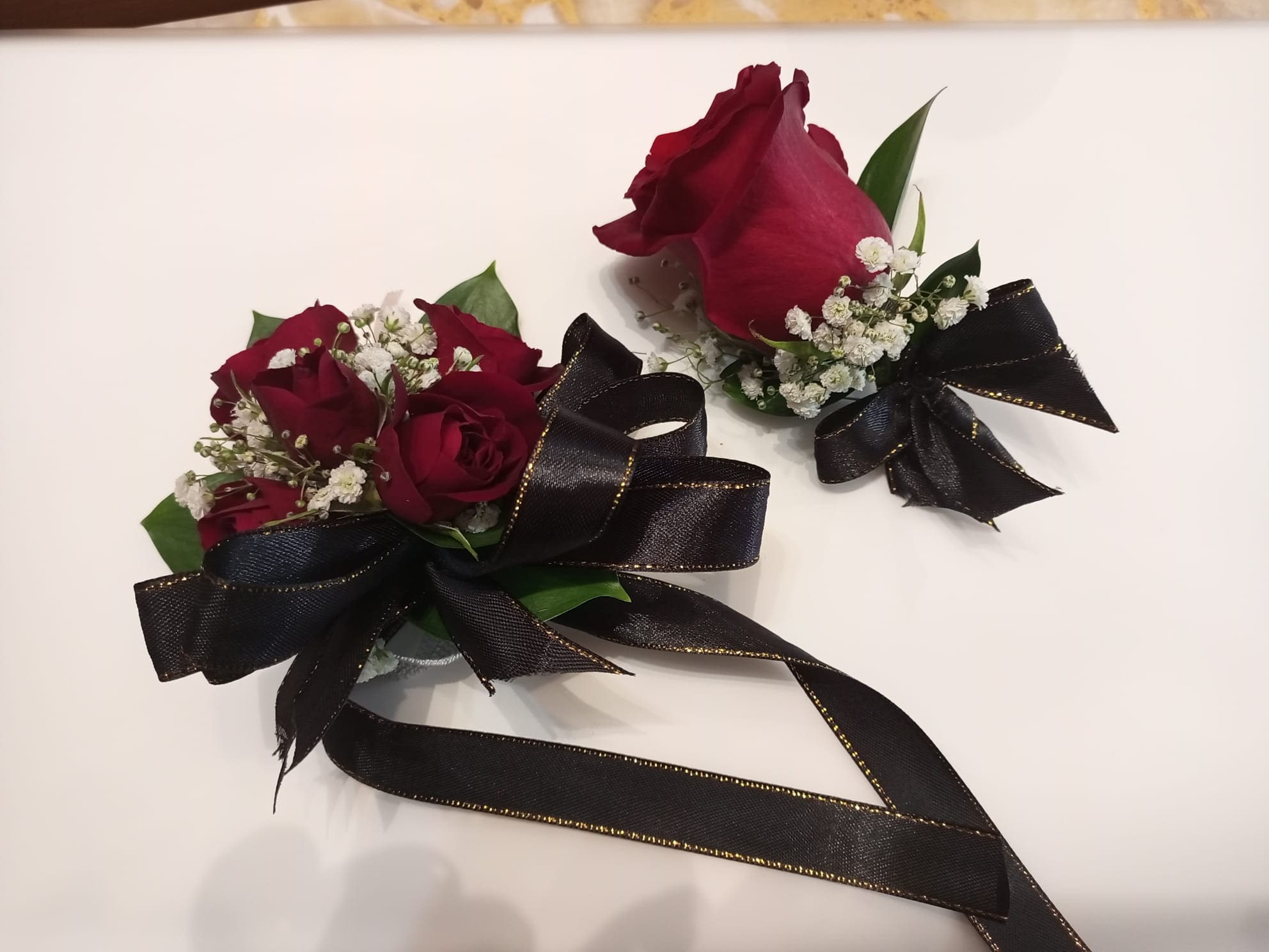 Black Ribbon Affair – Corsage and Boutonnière Set - A floral duo designed to stand out with elegance and sophistication. Black Ribbon Affair combines the intensity of red roses with the delicacy of white gypsum, wrapped in the distinction of a black satin bow with golden edges.  •	 The corsage, designed for the doll, is a floral jewel that enhances any outfit with its classic beauty.  •	 The boutonnière, elegant and discreet, adds a refined touch to the masculine or gala suit.