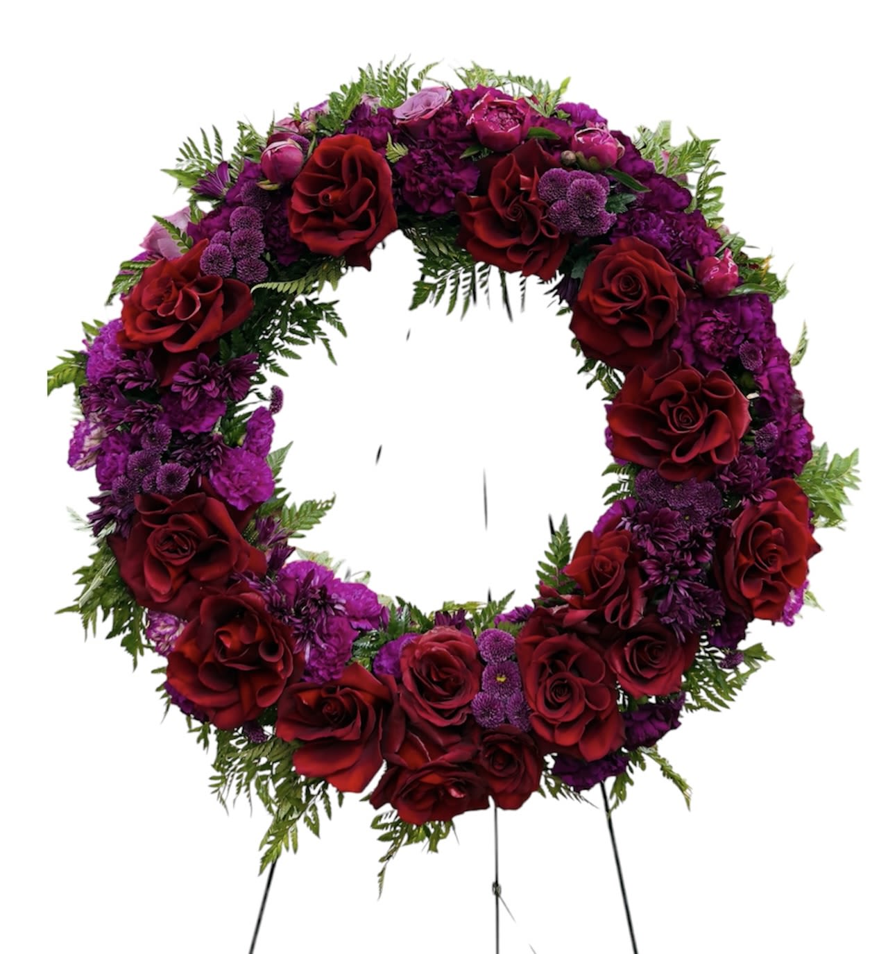 Red and purple Tribute Wreath - Deep crimson roses ring this large circular wreath, their velvety petals set against clustered purple chrysanthemums and vivid violet blooms for a dramatic sympathy arrangement that honors a life with quiet dignity. Designed as a formal memorial wreath for a funeral or graveside service, the stand-mounted presentation and open center create a respectful focal point that reads as both rich and timeless.  The composition pairs deep red roses, purple chrysanthemums, and complementary filler flowers, all softened by fern-like greenery for texture and contrast. Mounted on a sturdy wreath stand, the arrangement is sized to command presence at a service without overwhelming the space; the open center keeps the silhouette classic and recognizable from a distance.  Choose this tribute wreath when you want the person you're remembering to feel deeply honored-its traditional colors convey love, respect, and consolation. No special care is needed for the short-term display at a service; for longer placement, keep the wreath shaded and mist the flowers lightly to preserve freshness until the memorial concludes.