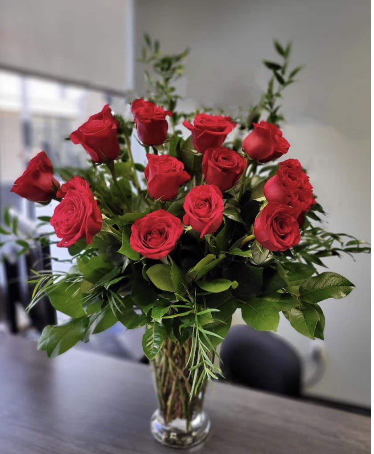 Rose Charm - A dozen deep red roses gathered into a full, rounded cluster above a clear glass vase-this Rose Charm arrangement reads as classic romance and timeless elegance, perfect for an anniversary or to say "I love you." The rich crimson petals and glossy green foliage create a lush, sculpted look that makes the recipient feel truly cherished.  Each bouquet features about twelve long-stemmed roses arranged tightly for a formal, traditional silhouette, with crisp white accents balancing the deep red and fresh greenery adding volume and contrast. The simple clear glass vase keeps the focus on the blooms and fits easily into home or office décor.  Choose this arrangement when you want someone to feel noticed and adored-ideal for anniversaries, Valentine's Day, or rekindling romance. Vase-ready and low-fuss, the roses arrive arranged for immediate display; keep fresh water, trim stems every few days, and remove spent blooms to extend their beauty.