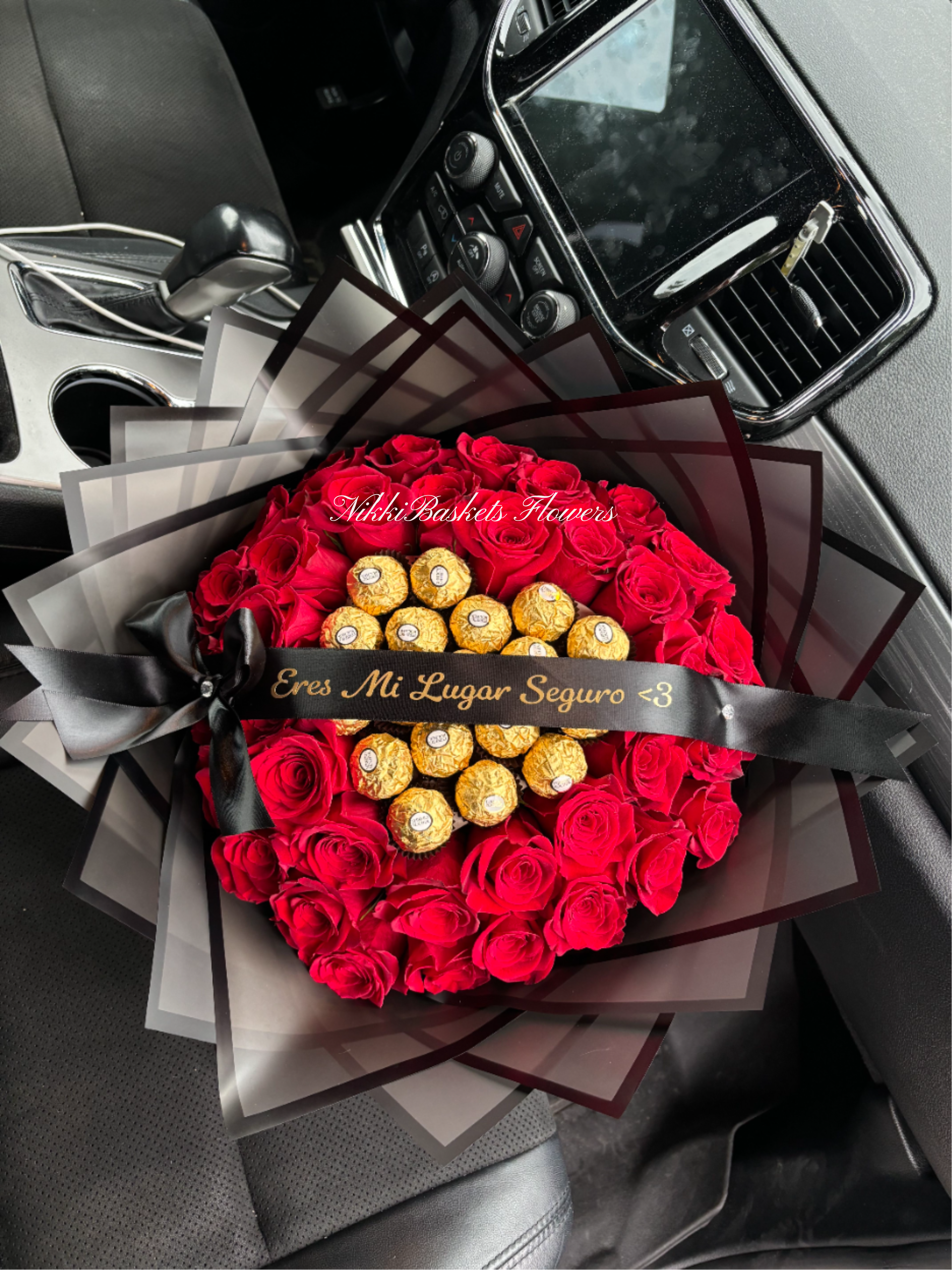 Round bouquet of red roses with gold-wrapped chocolates and black ribboned wrapping on a car seat.