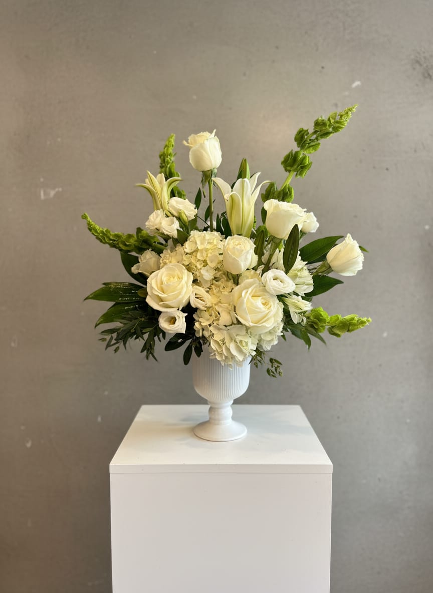 Graceful Tribute - Graceful Tribute is a timeless sympathy arrangement designed to honor life, express remembrance, and offer comfort during moments of loss.  This serene design features soft white roses, hydrangea, lilies, and seasonal textural blooms, thoughtfully arranged with lush greenery in a classic white pedestal vase. The palette is calm and refined—symbolizing peace, grace, and enduring love.  Created with a natural, garden-inspired shape, Graceful Tribute feels both elevated and heartfelt. It is an appropriate gesture for memorial services, celebrations of life, or sending comfort to a home when words are not enough. Each arrangement is handcrafted with care and intention in our studio.