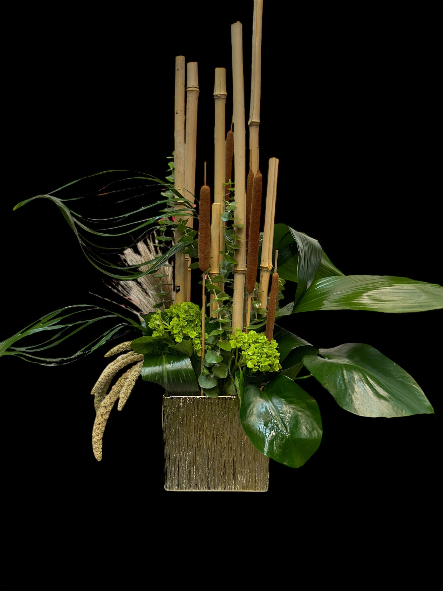 Tall tropical arrangement with bamboo stalks and broad green leaves in a gold vase