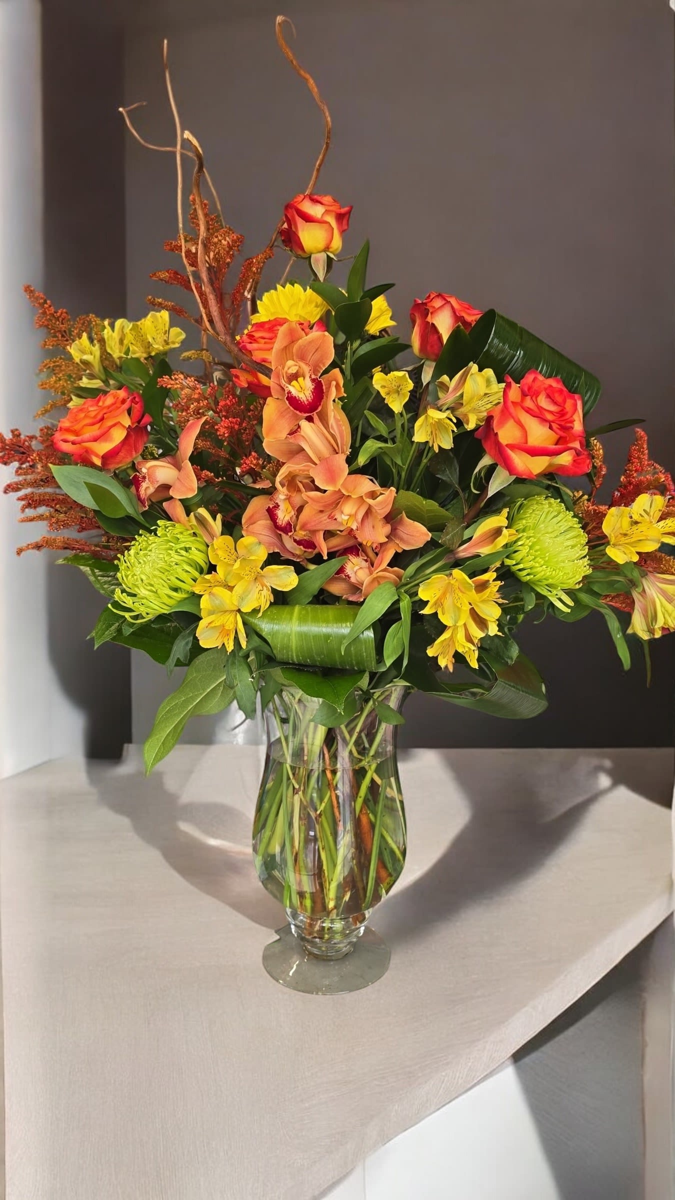 Mixed bouquet of orange roses, yellow flowers, and orchids in a glass vase