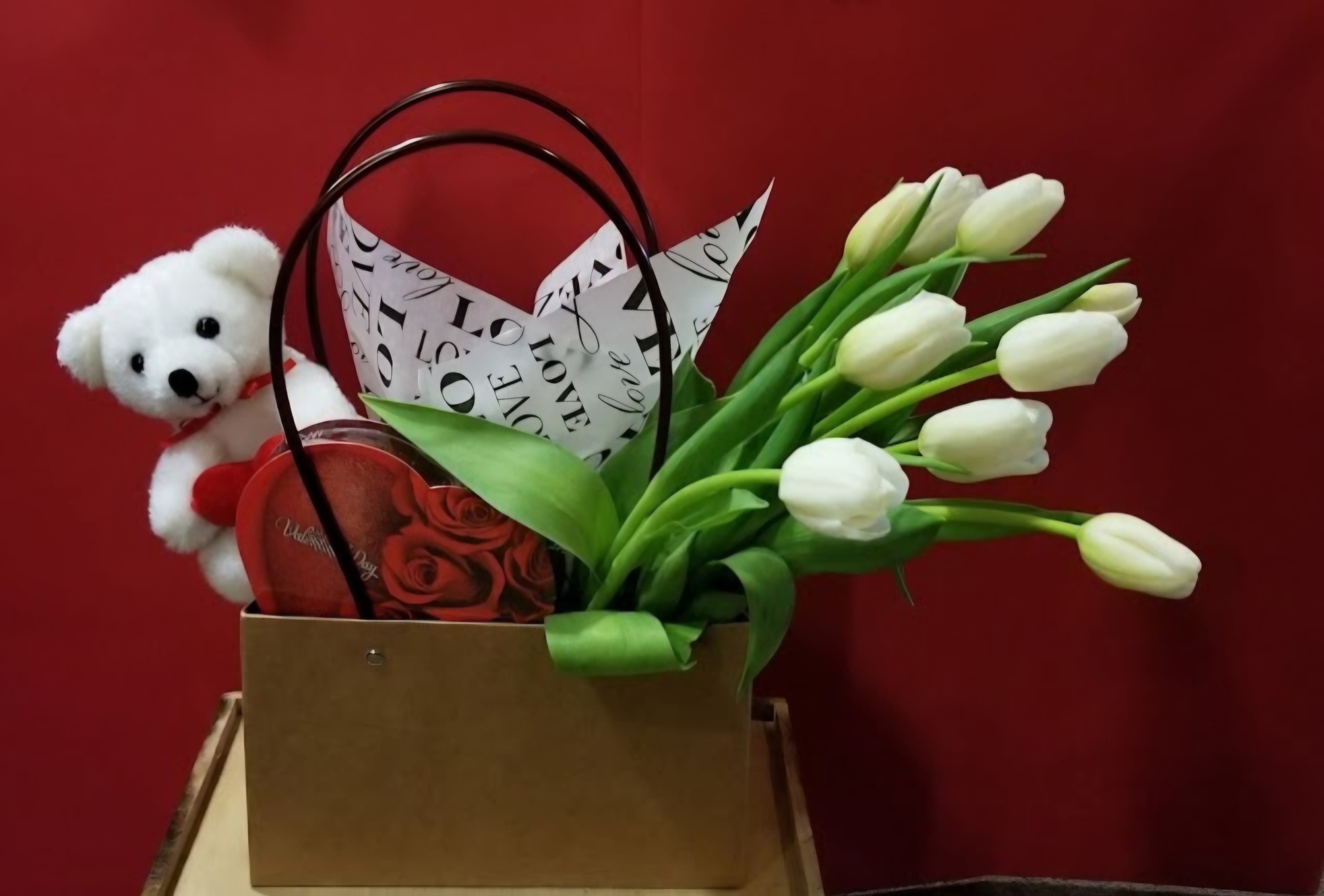 White tulips in a gift bag with a teddy bear and heart-shaped box