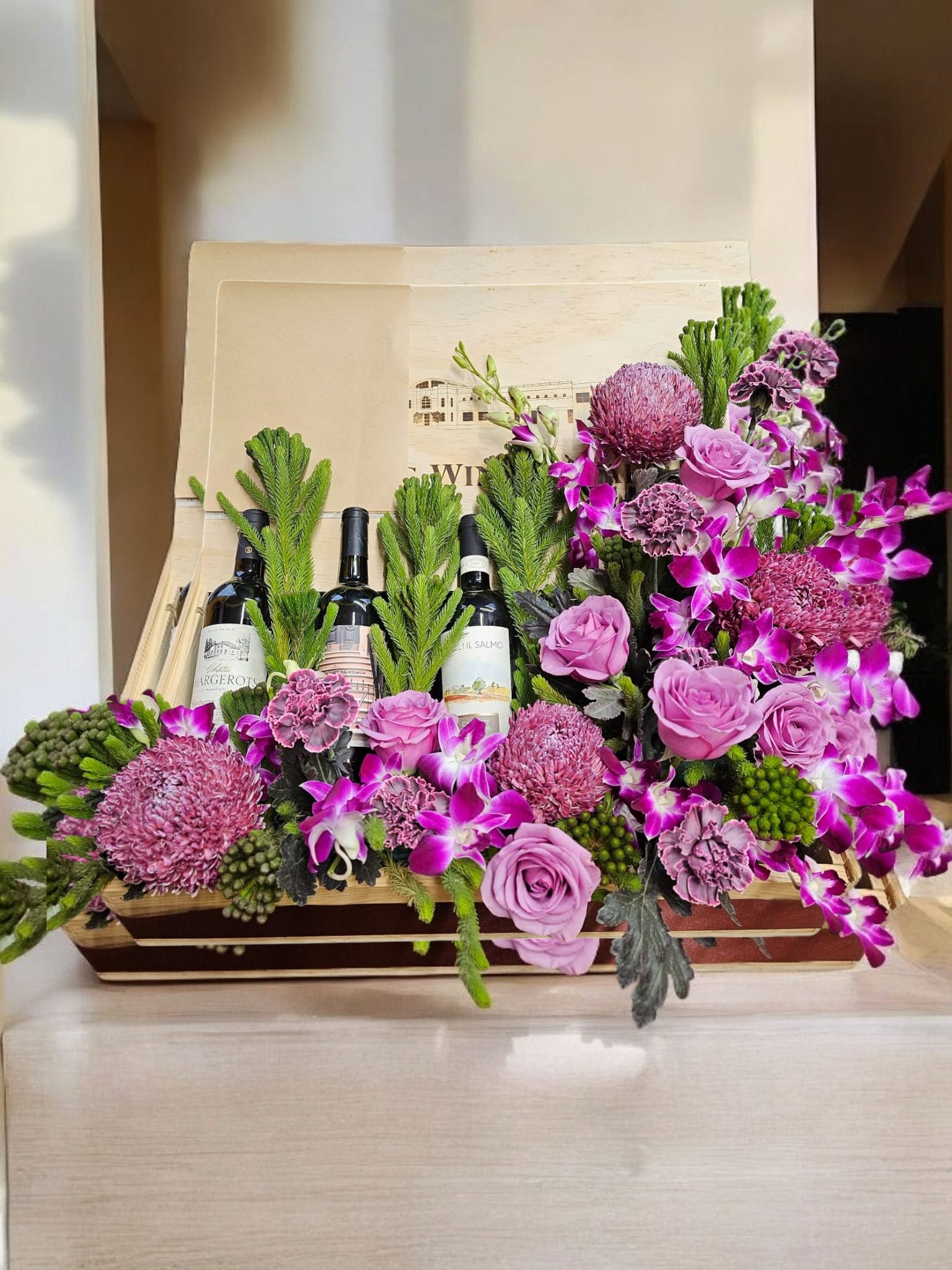 Pink and purple floral arrangement with wine bottles in a wooden box