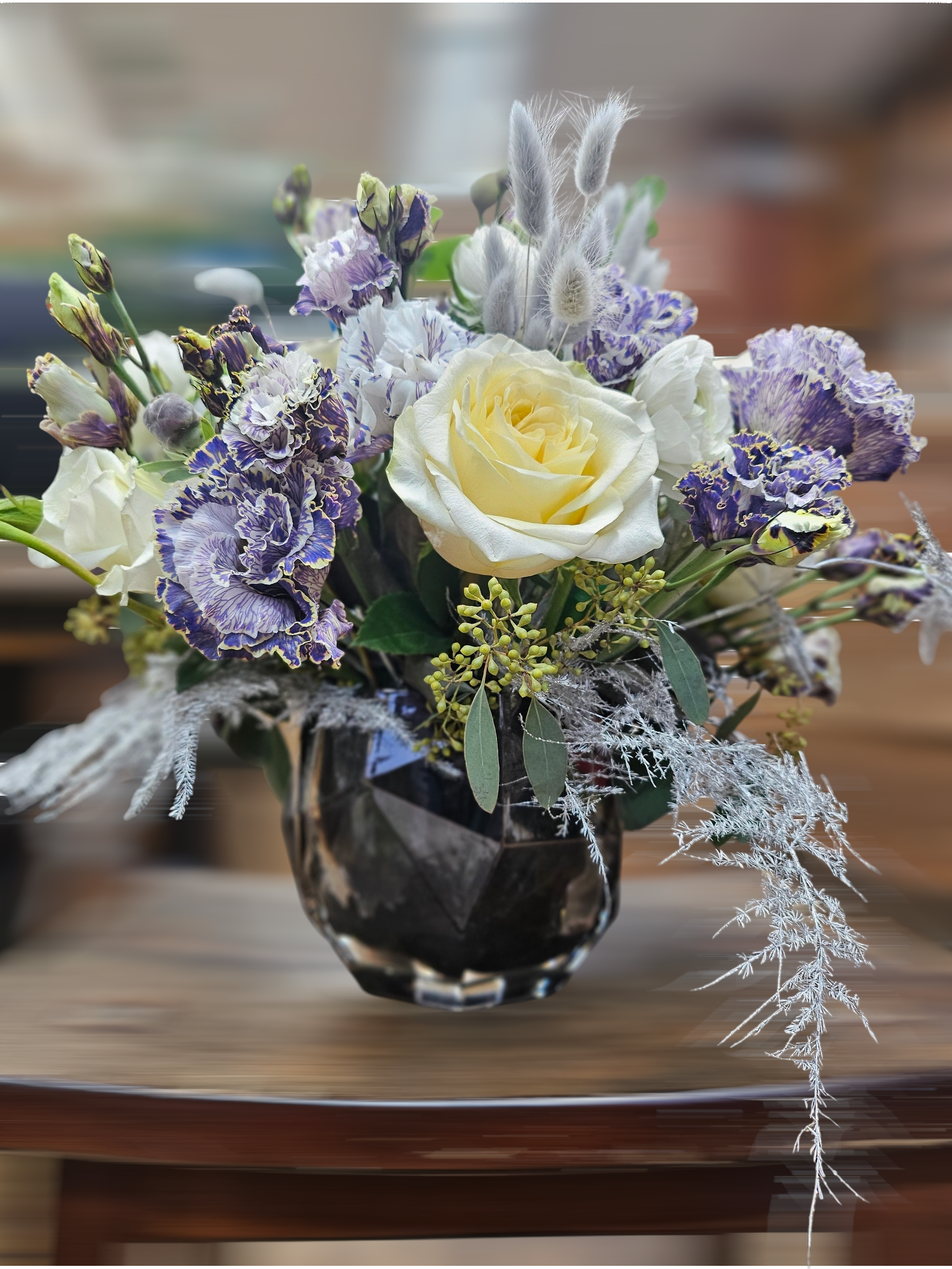 Short arrangement of cream roses and purple ruffled flowers in a dark glass vase