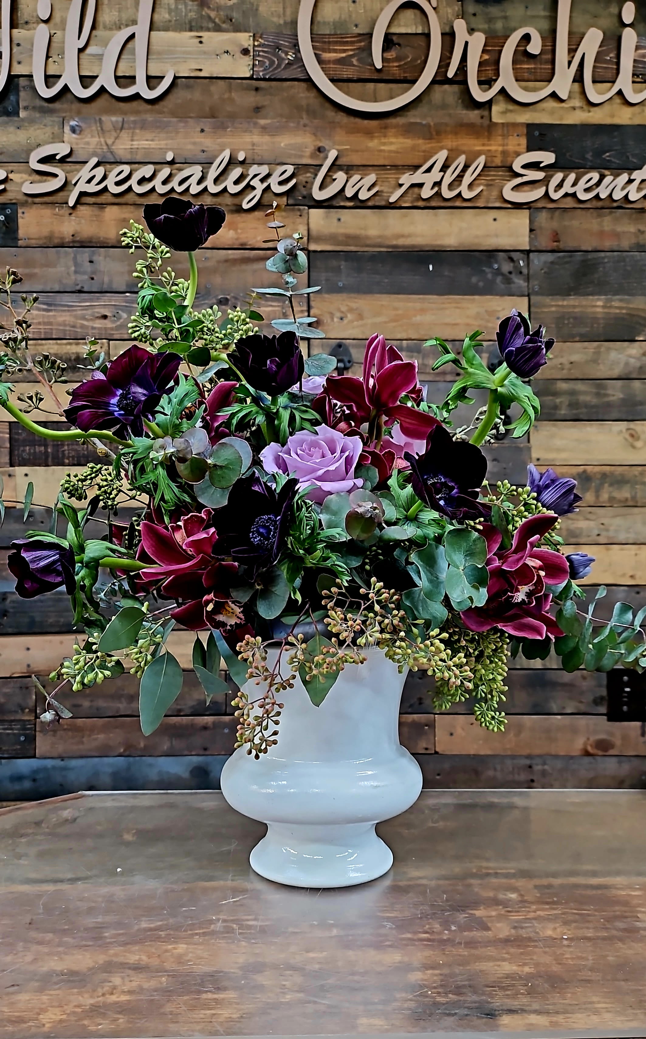 Purple and burgundy floral arrangement in a white pedestal vase