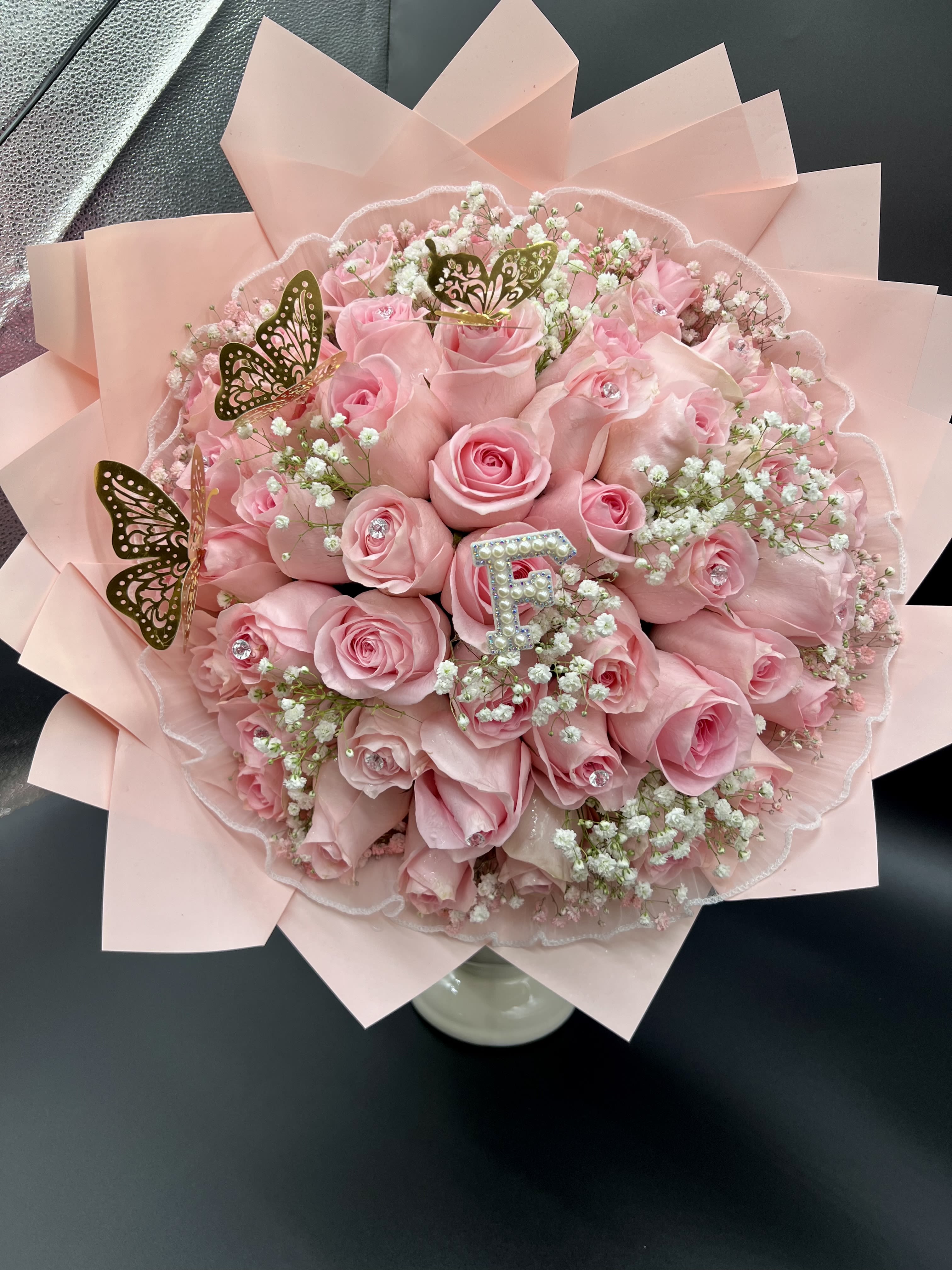 Round bouquet of pink roses and white filler flowers with gold butterfly accents wrapped in pale pink paper.