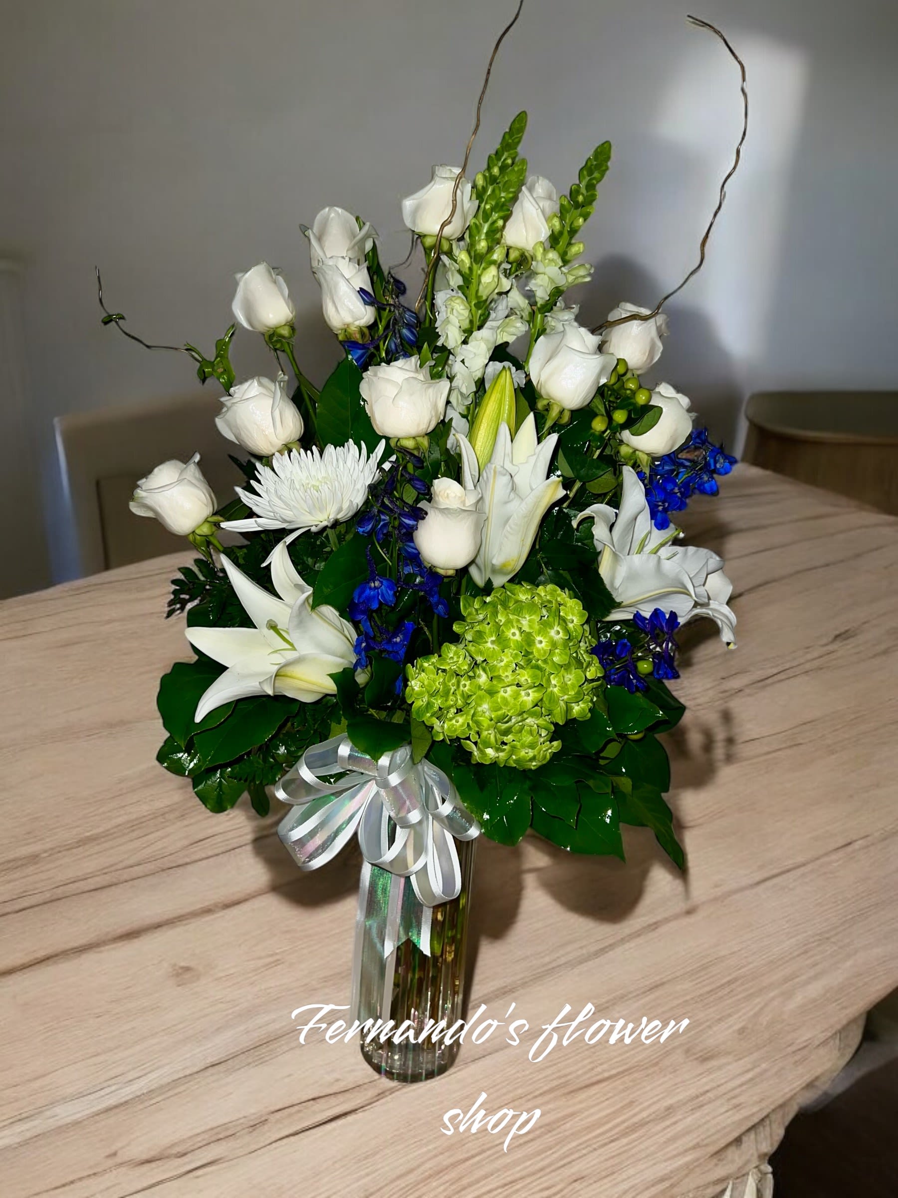 Tall arrangement of white roses and lilies with blue accents in a glass vase with a ribbon bow.