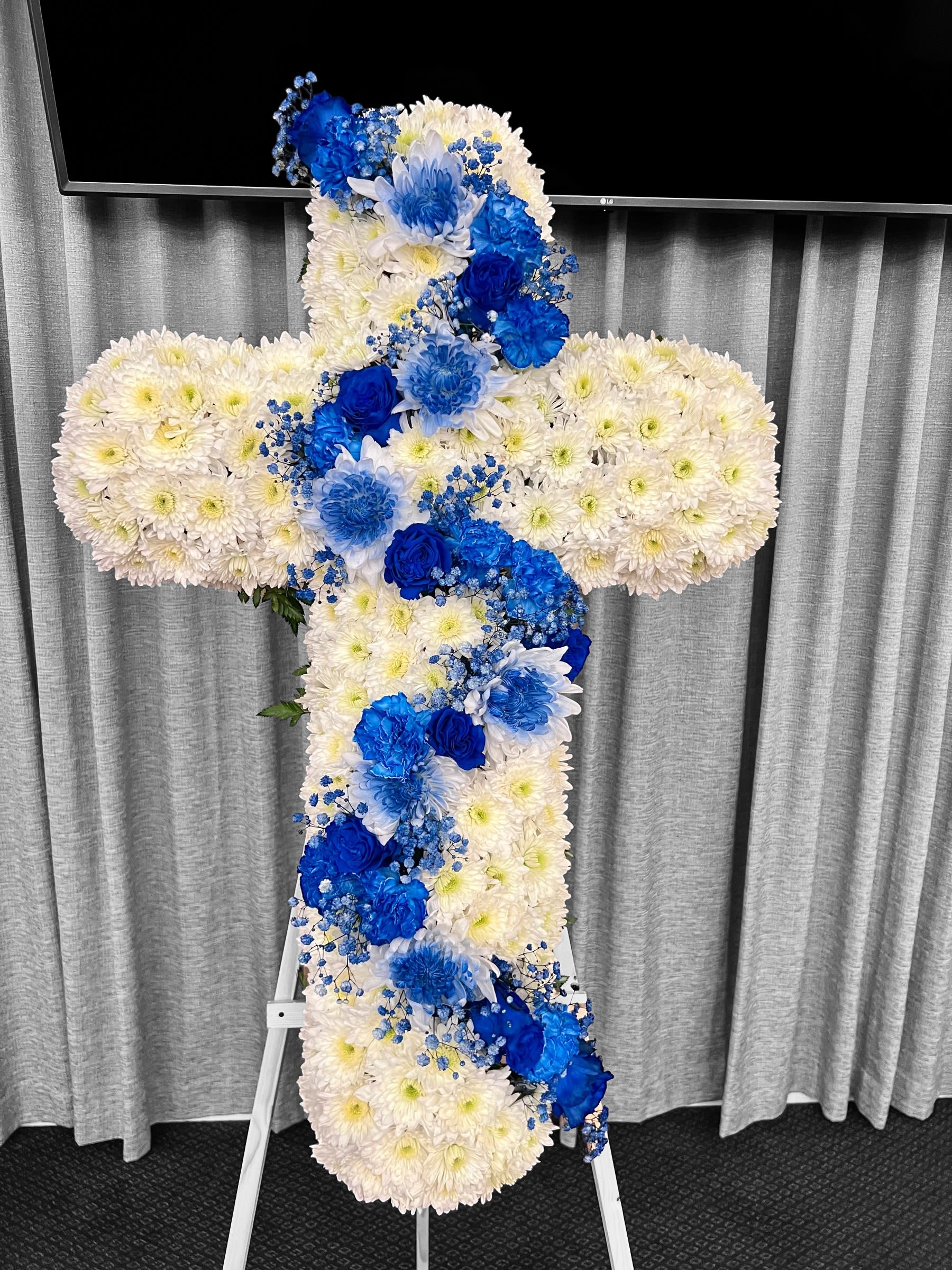 Standing cross of white flowers with cascading blue roses and blooms on an easel