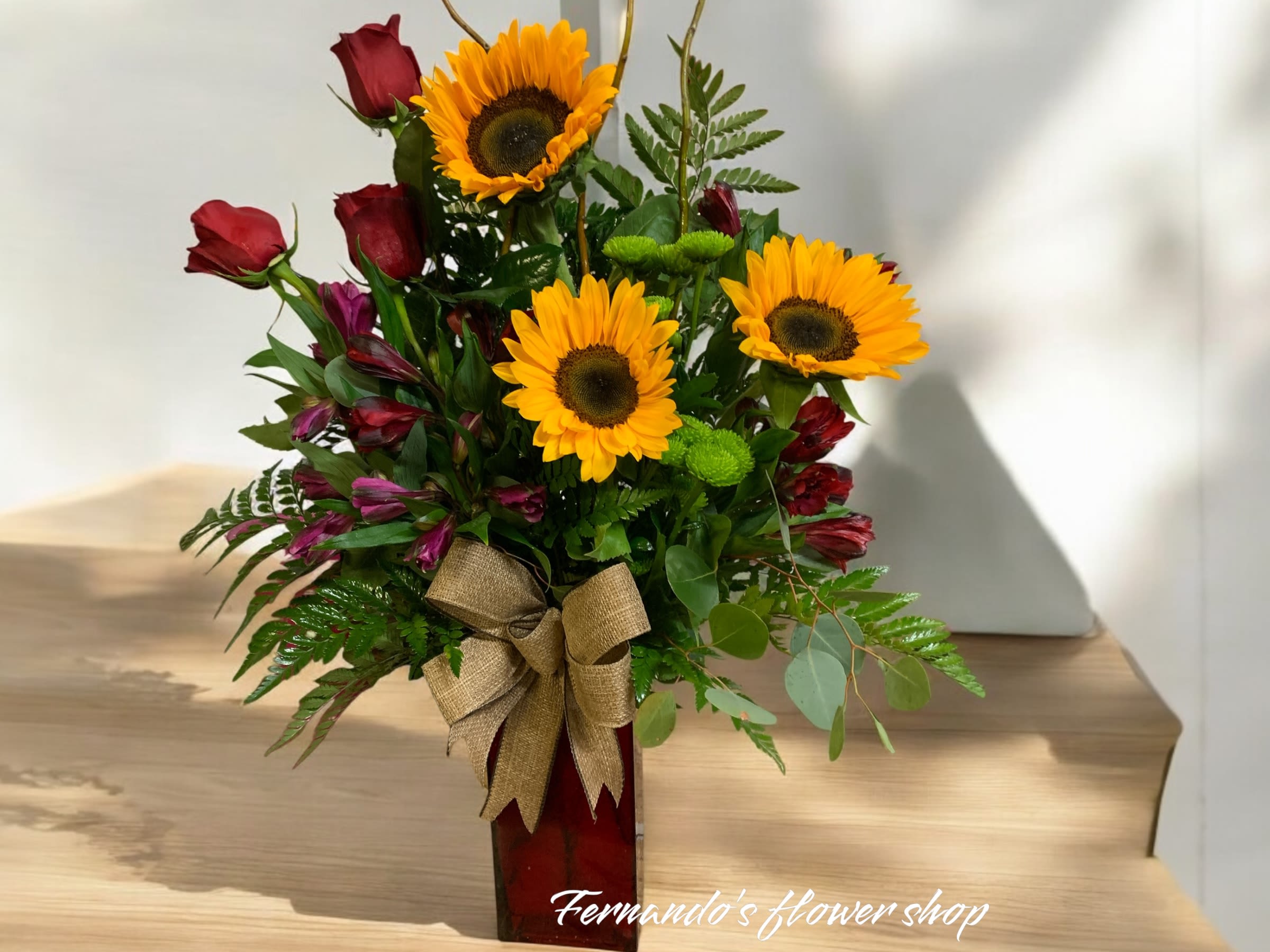 Arrangement of yellow sunflowers, red roses, and purple blooms in a red vase with a burlap bow