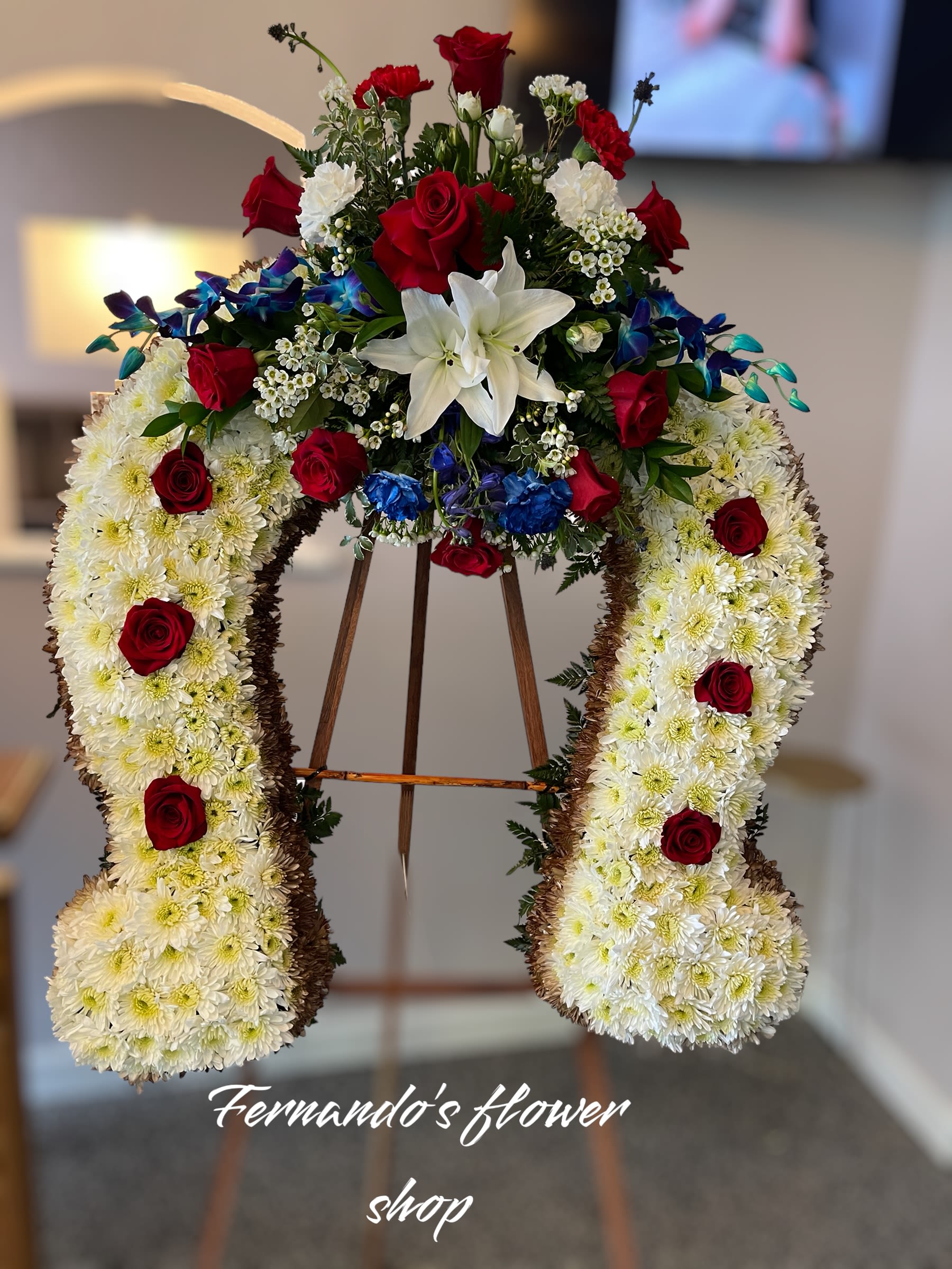 Horseshoe-shaped standing spray of white mums with red roses, lilies, and blue floral accents on an easel.
