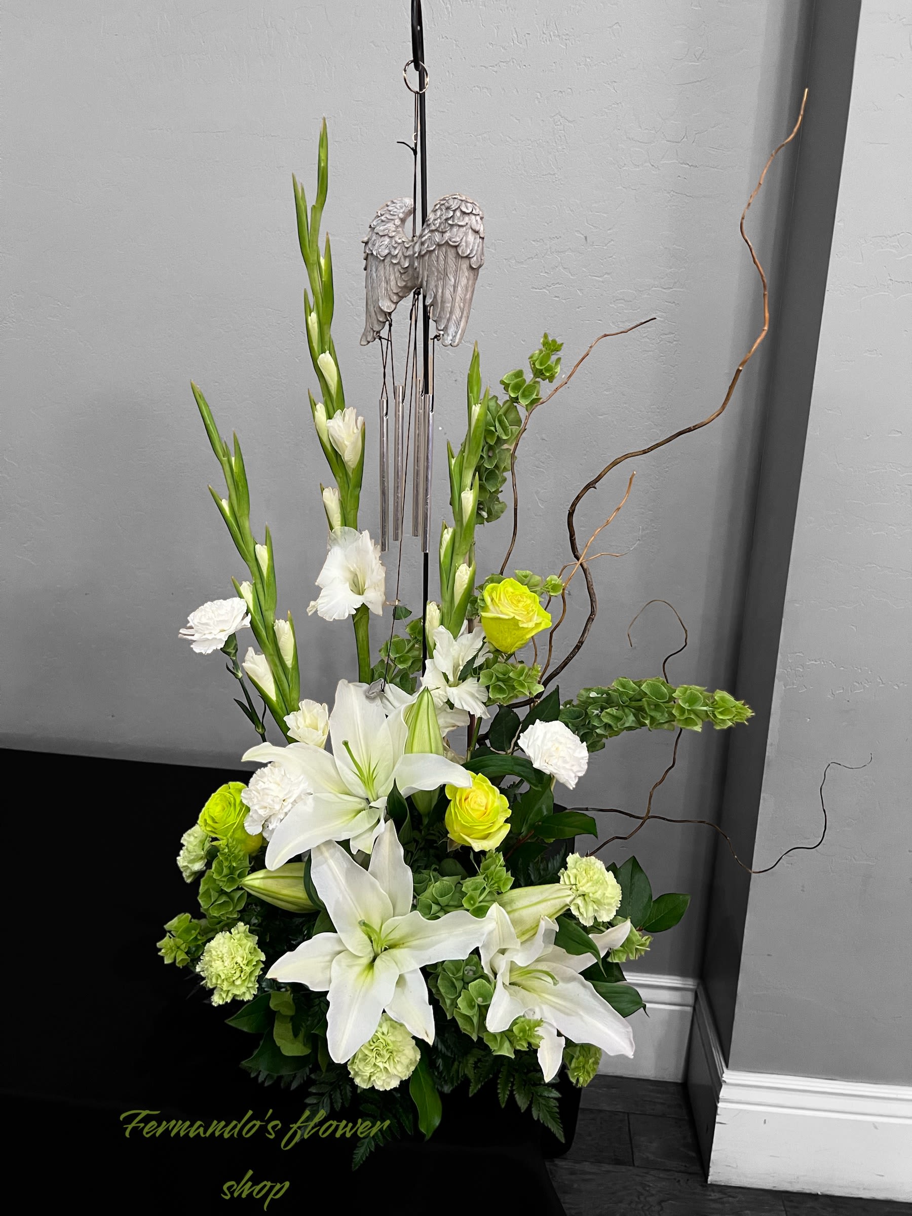 Wings and Bells - This elegant and heartfelt arrangement features pristine white lilies, carnations, and gladiolus, complemented by soft green roses, bells of Ireland, and lush greenery. Accentuated by natural curly willow branches, it adds a touch of rustic sophistication. At its center is a touching wind chime adorned with angel wings, symbolizing peace, remembrance, and eternal comfort. Perfect for honoring a loved one’s memory, this design is a serene and meaningful tribute, conveying love, grace, and solace during moments of reflection.