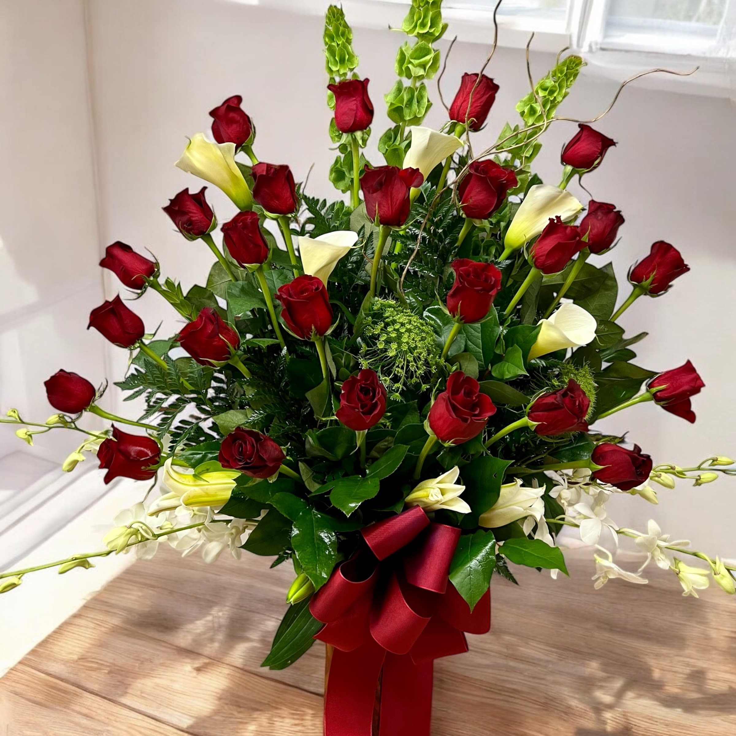 Tall arrangement of red roses, white calla lilies, and green bells of Ireland with a red bow.