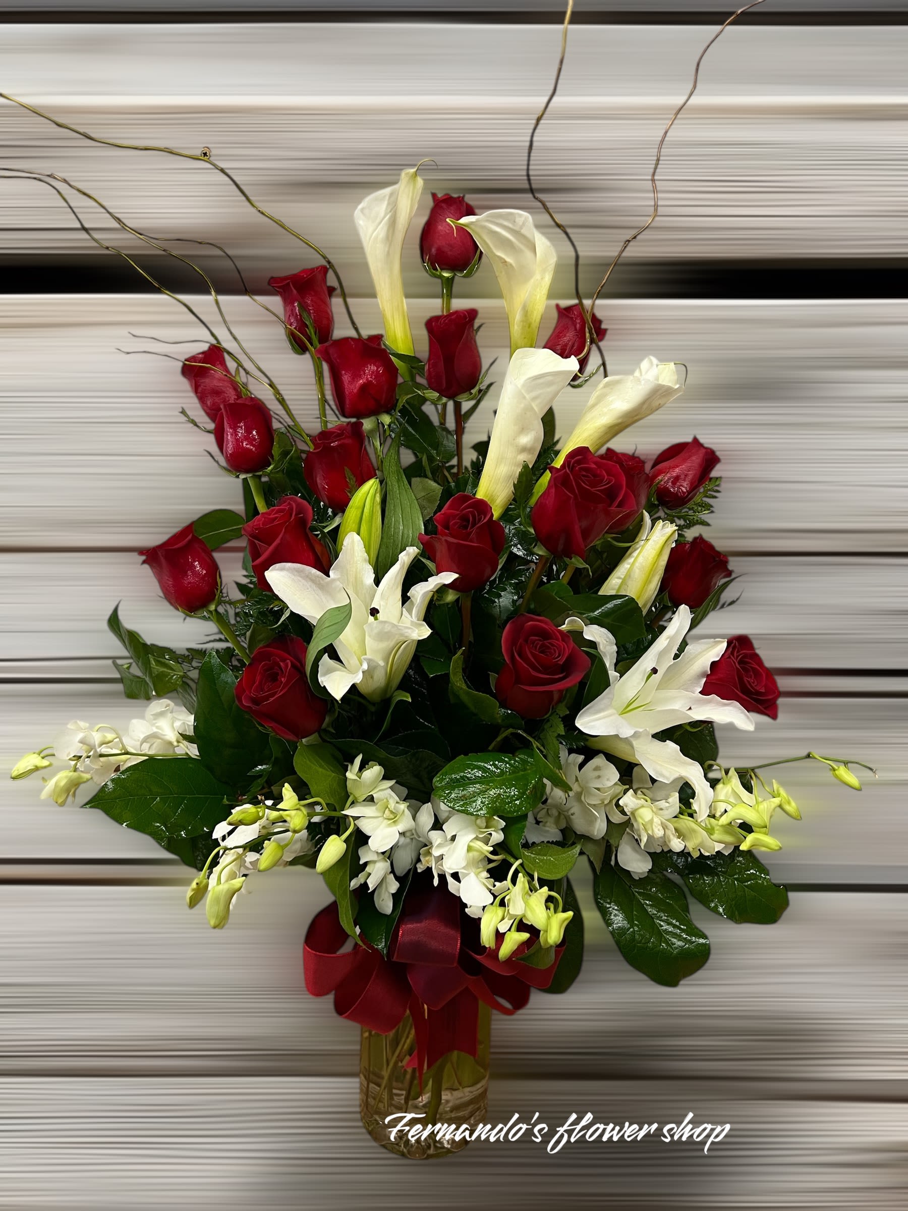Tall arrangement of red roses, white lilies, and calla lilies in a glass vase with a red ribbon