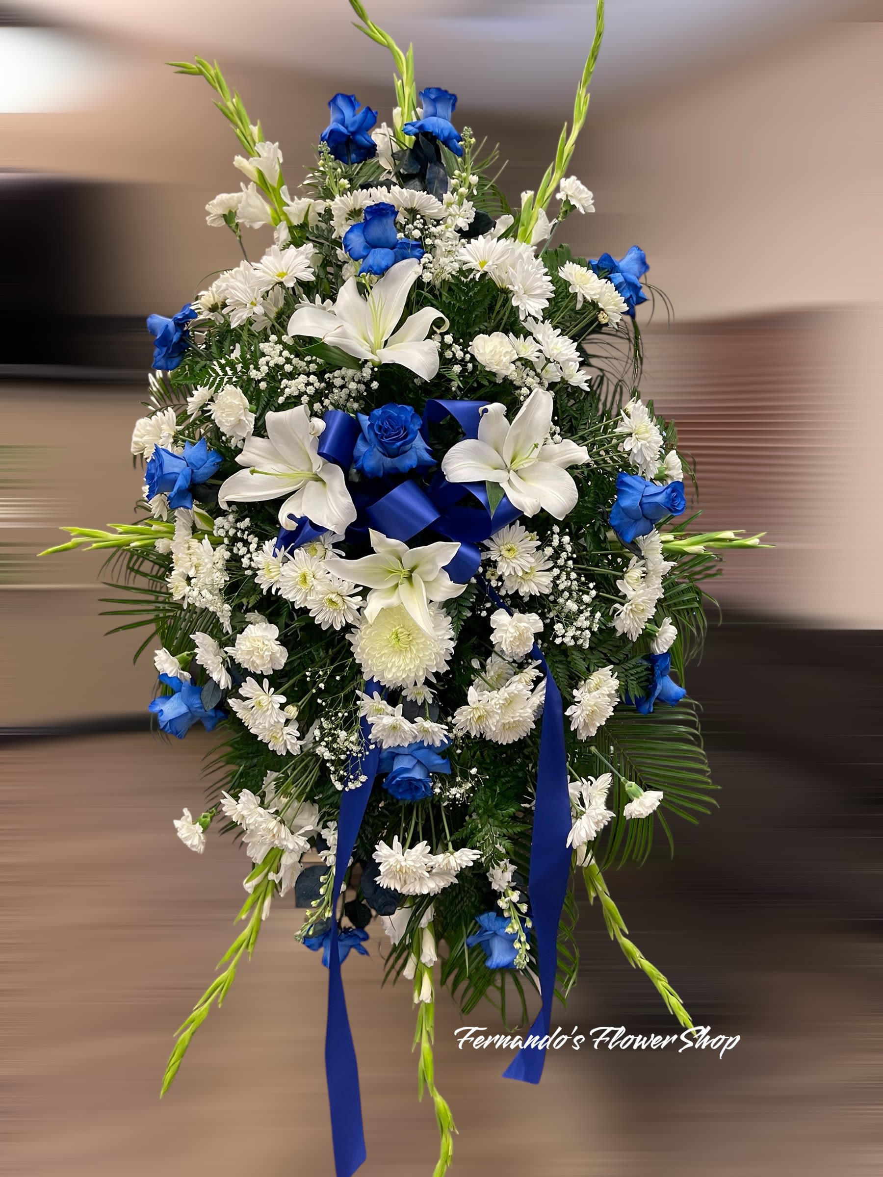 Tall standing spray of blue roses, white lilies, and white mums with blue ribbon on an easel.