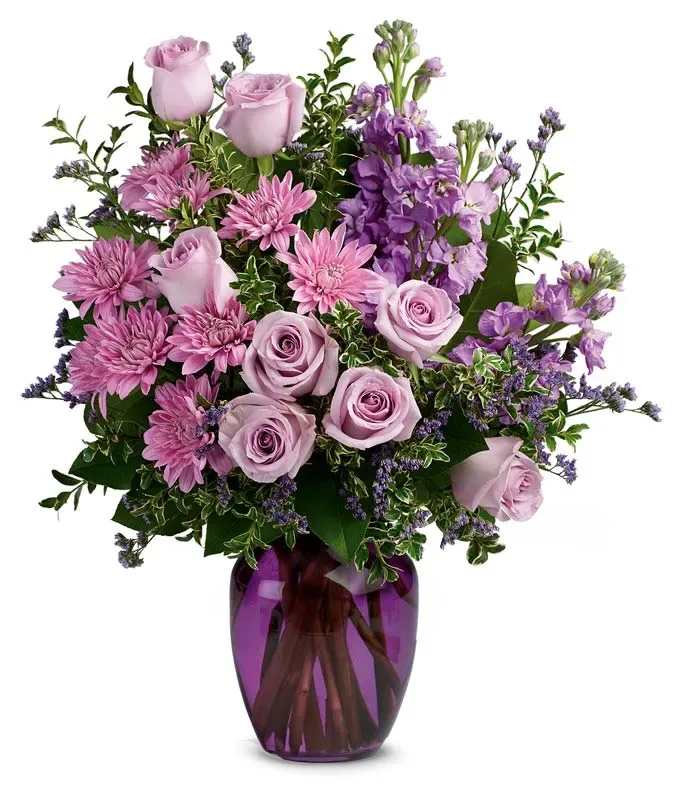 Lavender roses and pink mums arranged with purple blooms in a purple glass vase