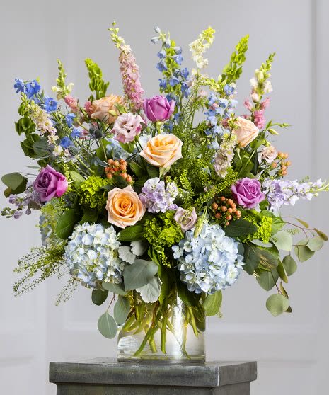 Mixed arrangement of lavender and peach roses, blue hydrangeas, and tall delphinium in a clear glass vase